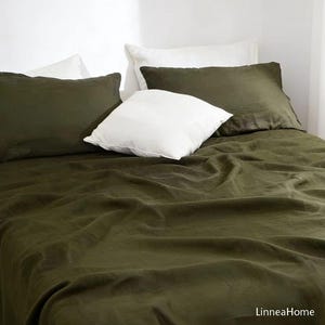 May include: A bed with olive green linen bedding, including a fitted sheet and pillowcases. Two olive green pillows and two white pillows are arranged on the bed. The bedding is made of a soft, wrinkled fabric. The brand name "LinneaHome" is visible in the bottom right corner.