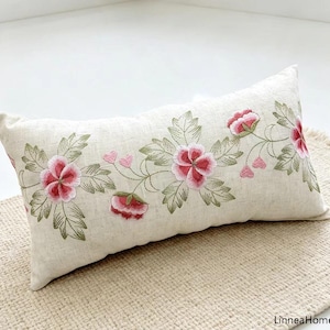 Puede incluir: Un cojín rectangular de tela beige claro con un patrón de enredadera bordado horizontalmente. El diseño incluye flores rosas y rojas con hojas verdes y pequeños detalles de corazones rosas. El texto LinneaHome es visible.