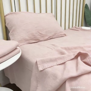 May include: A bed with a light pink linen sheet set. The set includes a pillow, a fitted sheet, and a flat sheet. The bed frame is gold. The sheets are neatly arranged, with soft folds and a relaxed appearance. The overall aesthetic is minimalist and inviting.
