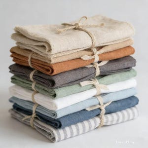 May include: A stack of folded linen napkins in various colors, including beige, brown, gray, green, and blue. Each stack is tied with twine. The bottom napkin has a striped pattern.