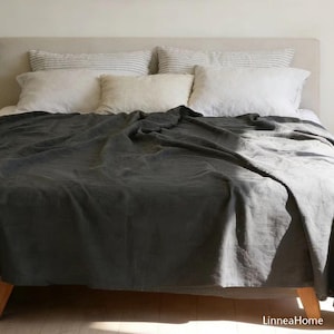 May include: A dark gray linen bedspread draped over a bed, with white and striped pillows. The bed has a light-colored headboard and wooden legs. The bedspread is made of a soft, textured fabric.