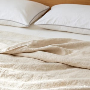May include: A beige linen bedspread with a visible woven texture draped over a bed. Two white pillows with thin grey piping are positioned at the head of the bed. The text "LuneaHome" is visible in the bottom right corner of the image.