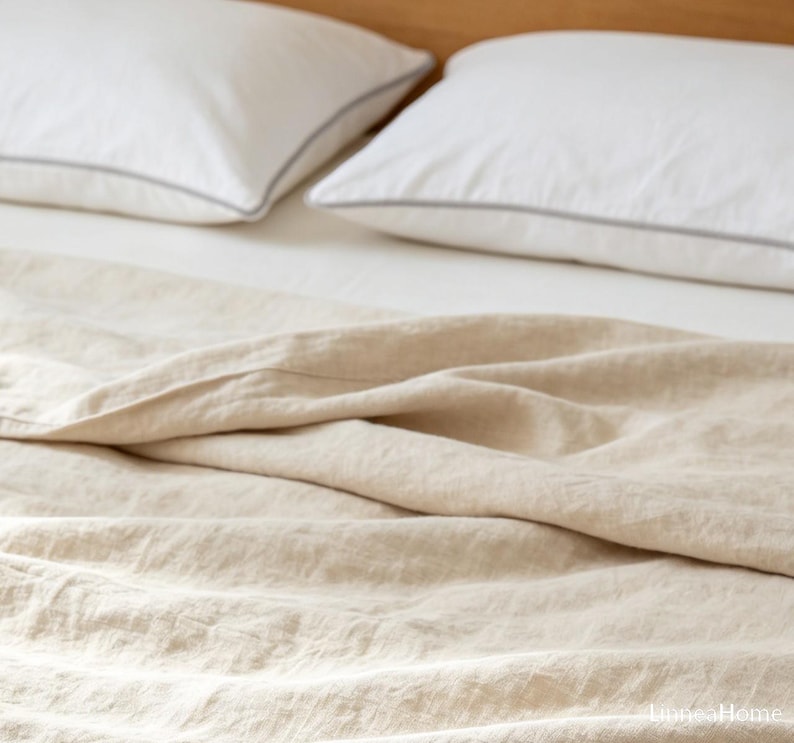 May include: A beige linen bedspread with a visible woven texture draped over a bed. Two white pillows with thin grey piping are positioned at the head of the bed. The text "LuneaHome" is visible in the bottom right corner of the image.