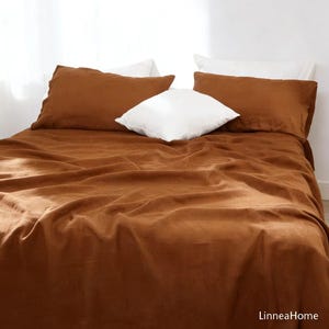 May include: A bed covered with a rust-colored linen sheet set. Two rust-colored pillowcases and one white pillow are visible. The bedding is arranged on a bed in front of a white wall. The text "LinneaHome" is in the bottom right corner.