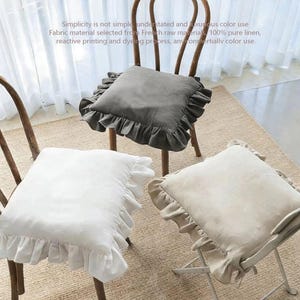 May include: Three chair cushions in different colors: gray, white, and beige. Each cushion has a ruffled edge. The cushions are on wooden and metal chairs, placed on a neutral-colored rug. The text on the image says "Simplicity is not simple, understated and luxurious color use."