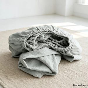 May include: A light gray fitted sheet with an elasticized edge, featuring a soft, textured fabric surface. The sheet is gathered in a circular shape, displaying the flexible, stretchy border designed to secure the bedding around a mattress.