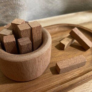 May include: A wooden bowl filled with small, rectangular wooden blocks. Several blocks are scattered on a wooden tray. The items are made of a light brown wood, creating a natural and rustic aesthetic.