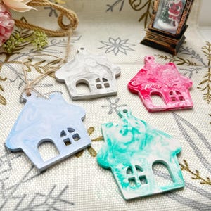 May include: Four house-shaped ornaments with a marbled effect in blue, white, pink, and teal. Each has a cutout window and a small hole for hanging. The ornaments are on a decorative surface, suggesting a festive theme, possibly Christmas.