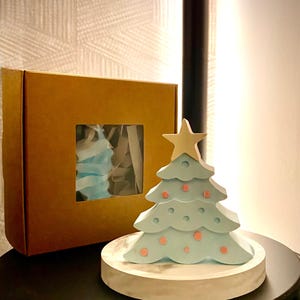 May include: A light blue, tiered Christmas tree figurine with a pale yellow star topper and small pink circular accents. The tree sits on a round, white marble-patterned base next to a square, gold-coloured cardboard gift box with a clear window.