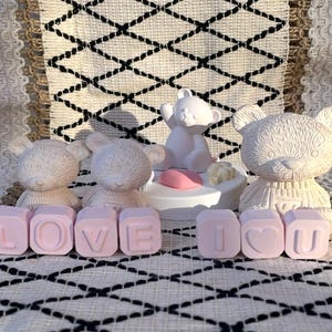 May include: A display of decorative items featuring off-white teddy bear figurines and pink letter blocks spelling "LOVE I U". The bears are in different poses and sizes, with one on a pedestal. The items are arranged on a patterned surface.