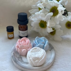 May include: Three rose-shaped soaps in pink, blue, and white sit on a marble tray. Two small amber bottles and a larger essential oil bottle with a black cap are next to the tray. White daisies are in the background.