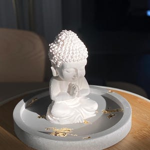 May include: A white Buddha statue in a meditative pose, seated on a grey circular tray with gold leaf accents. The statue has a textured head and is placed on a wooden surface.
