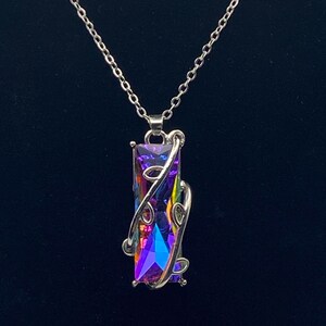 Puede incluir: Un collar plateado con un colgante rectangular iridiscente. El colgante presenta una vibrante mezcla de tonos morados, azules y verdes, con un diseño decorativo de remolino plateado. La cadena es delicada, sobre un fondo oscuro.