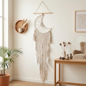 May include: A cream-colored macrame wall hanging in a crescent moon shape. The boho-style decor features a wooden dowel, intricate knotting, and dangling fringe with feathers and beads. It hangs on a white wall, adding a touch of bohemian charm to any space.
