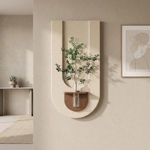 Könnte beinhalten: Ein beigefarbener Wandtopf mit einem Holzregal und einer klaren Glasvase mit einer grünen Pflanze. Der Pflanztopf hat ein geschwungenes Design mit vertikalen Linien. Ein gerahmter Kunstdruck ist rechts zu sehen.