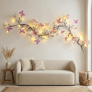 Peut inclure: Décoration murale représentant une branche sombre ornée de papillons roses et violets, illuminée par de petites lumières chaudes. L'ensemble est placé au-dessus d'un canapé beige, ajoutant une touche décorative à la pièce.