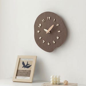 May include: A brown wooden wall clock with a unique, organic shape. The clock features light-colored numbers and hands, with a natural wood grain finish. The clock is mounted on a white wall. A framed print and decorative items are in the foreground.