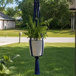 May include: A white ceramic plant pot with a textured surface held by a deep blue macrame hanger. The hanger features intricate knotted cord patterns and terminates in a long, decorative tassel at the bottom, suspending the plant in the air.