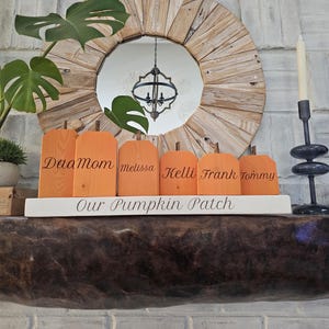 Può includere: Un set di sei blocchi di legno arancioni a forma di zucca con steli marroni, ognuno inciso con un nome: Daa, Mom, melissa, Kelli, Frank e Tommy. I blocchi poggiano su una base rettangolare bianca con la scritta Our Pumpkin Patch in corsivo.