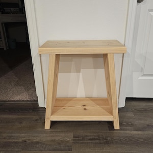 Könnte beinhalten: Kleiner, unbehandelter Beistelltisch aus Holz mit rechteckiger Oberseite und unterem Regal. Die Beine sind abgewinkelt, und das Holz hat eine natürliche, helle Farbe. Der Tisch ist für den Innenbereich konzipiert.