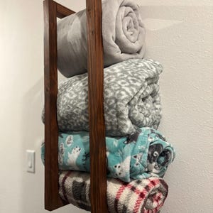 May include: A vertical wall-mounted wooden storage rack with a dark brown finish, holding four rolled blankets. The blankets include a solid light grey, a grey and white leopard print, a light blue cat print, and a red, grey, and white plaid pattern.