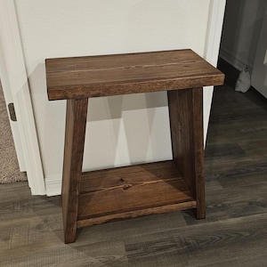 May include: A small, rectangular wooden side table with a dark brown stain. The table has a top surface, a lower shelf, and four angled legs. The wood grain is visible, and the table is sturdy.
