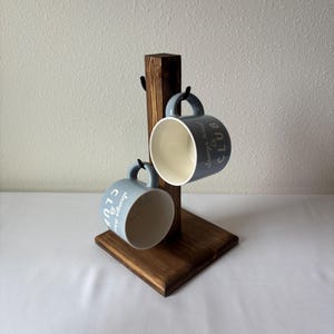May include: A dark brown wooden mug tree stand with a square base and a vertical post featuring two black metal hooks. Two light blue ceramic mugs with white interiors are hanging from the hooks, each printed with the text "always tired CLUB".