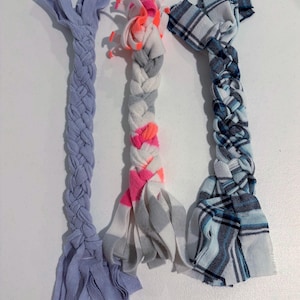 Può includere: Tre giochi di corda in tessuto intrecciato. Il gioco a sinistra è di colore viola chiaro a tinta unita. Quello centrale presenta strisce di tessuto bianco, grigio e rosa neon. Quello a destra è in tessuto a quadri blu, bianco e nero. Ogni gioco ha le estremità sfilacciate.