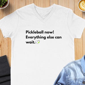 May include: White V-neck t-shirt with the text "Pickleball now! Everything else can wait." in black, with a green pickleball and heart graphic. The shirt is displayed on a wooden surface.