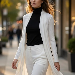 May include: A long-sleeved white open-front duster cardigan worn over a black turtleneck sweater and paired with matching white tailored trousers. The ensemble features a clean, layered look with a solid color palette and smooth fabric texture.