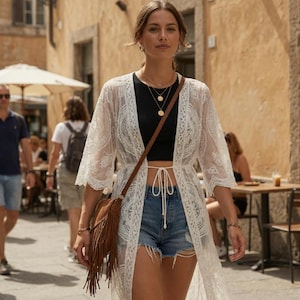 May include: A woman wearing a white lace kimono, black top, and denim shorts walks down a street. She carries a brown fringed bag and wears gold necklaces. The background features a European street scene with cafes and pedestrians.