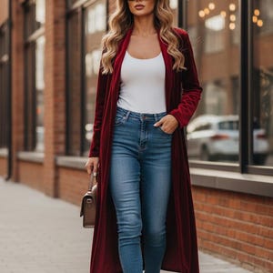 May include: A woman wearing a long, burgundy velvet coat over a white top and blue jeans. She is also wearing black sunglasses and black heels, carrying a brown purse. The background features a brick building and large windows.