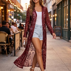 May include: A long-sleeved, floor-length burgundy duster cardigan featuring an intricate, sheer lace overlay with shimmering sequin embellishments. The garment is worn over a plain white tank top and light-wash denim shorts with frayed hems.