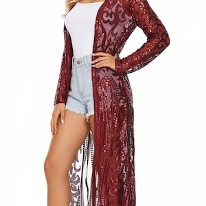 May include: A long, burgundy sequined cardigan with an open front and long sleeves. The cardigan features an intricate pattern and is worn over a white top and denim shorts. The model is wearing white high-heeled sandals.