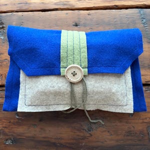 Può includere: Una custodia rettangolare in feltro con una patta blu vivace e un corpo grigio chiaro. Una striscia verticale di feltro verde salvia corre lungo il centro, fissata da un bottone in legno chiaro con un sottile spago verde avvolto attorno.