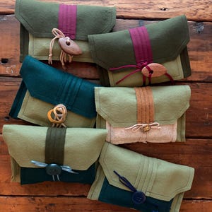 May include: Six rectangular felt pouches in various shades of green, olive, and beige. Each pouch features a flap closure secured with a unique button, including wooden, metallic, and plastic styles, tied with thin cord or string accents.