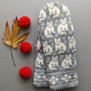 Puede incluir: Un par de mitones de punto gris y blanco con un patrón de gato repetido. Los mitones están junto a una hoja seca y tres pompones rojos sobre una superficie gris. Un accesorio de invierno acogedor.