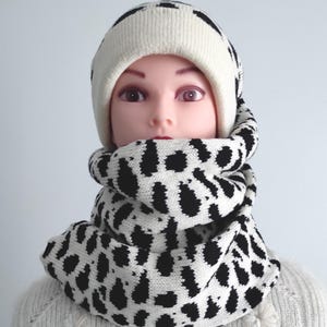 May include: A knitted winter set featuring a white beanie with black stripes and a matching cowl scarf. The scarf and hat have a black and white spotted pattern. The mannequin is wearing a white jumper with floral embellishments.
