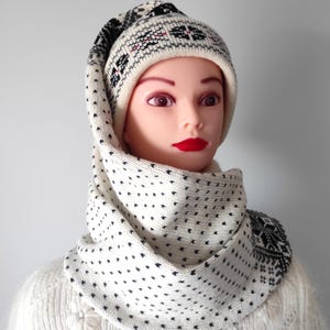May include: A knitted hat and cowl set in cream and black. The hat features a geometric pattern in black, red, and gray. The cowl is dotted with small black shapes. The set is displayed on a mannequin.