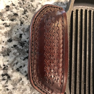 May include: A close-up of a brown leather handle cover with a woven design. The cover is attached to a cast iron grill pan.
