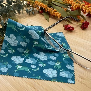 May include: A teal blue microfiber cleaning cloth with a repeating pattern of white clouds and green dragons. A pair of silver-rimmed glasses is lying on top of the cloth.