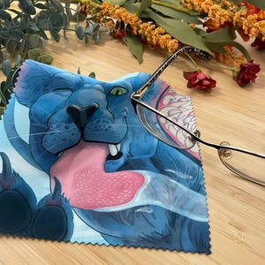 May include: A blue microfiber cleaning cloth with a cartoon illustration of a blue cat with a pink tongue sticking out. The cat has a white background and a black outline. The cloth is folded in half and has a white border.