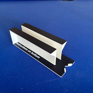 May include: A black and white plastic stand featuring two parallel vertical walls connected to a flat base. The side of the stand displays the white text "Instructions on bottom" printed on a black surface, with a V-shaped cutout at one end.