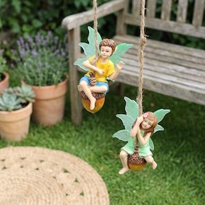 May include: Two fairy figurines swing from a rope. One fairy wears a yellow shirt and blue shorts, the other wears a green dress. Both are seated on acorn swings with green translucent wings. The background includes a wooden bench and potted plants.