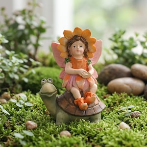 May include: A decorative figurine featuring a small fairy with brown hair and a sunflower-shaped hat, wearing an orange and pink dress with wings, sitting atop a smiling turtle with a brown shell, crafted with detailed textures and colours.