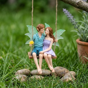 Peut inclure: Un couple de fées miniatures sur une balançoire. Le garçon porte une chemise et un short bleus, tenant une fleur jaune. La fille porte une robe violette. Les deux ont des ailes vertes et sont assis sur une balançoire en bois suspendue par de la ficelle.