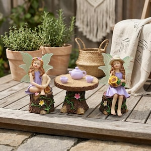 May include: A whimsical fairy garden scene featuring two fairy figurines in purple dresses with flower crowns and translucent wings. A small wooden table with a teapot and teacups sits between them. The fairies are seated on tree stump bases.