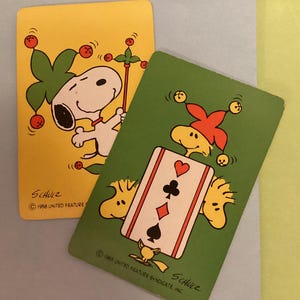 May include: Two vintage Snoopy playing cards. One card is yellow with Snoopy dressed as a jester. The other card is green with a card showing the suit of hearts, clubs, and diamonds. The cards are from 1958 and 1965.