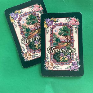Op de afbeelding: Twee rechthoekige speelkaarten met een groene rand en het woord "Tennessee" in sierlijke letters. De kaarten tonen een landschap met bomen, een weg en bloemen, omlijst door een bloemenrand.