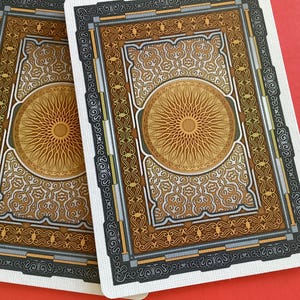 May include: Two playing cards with ornate designs. The backs feature a central sunburst pattern in gold, surrounded by intricate borders in shades of brown, gray, and gold. The cards are set against a red background.